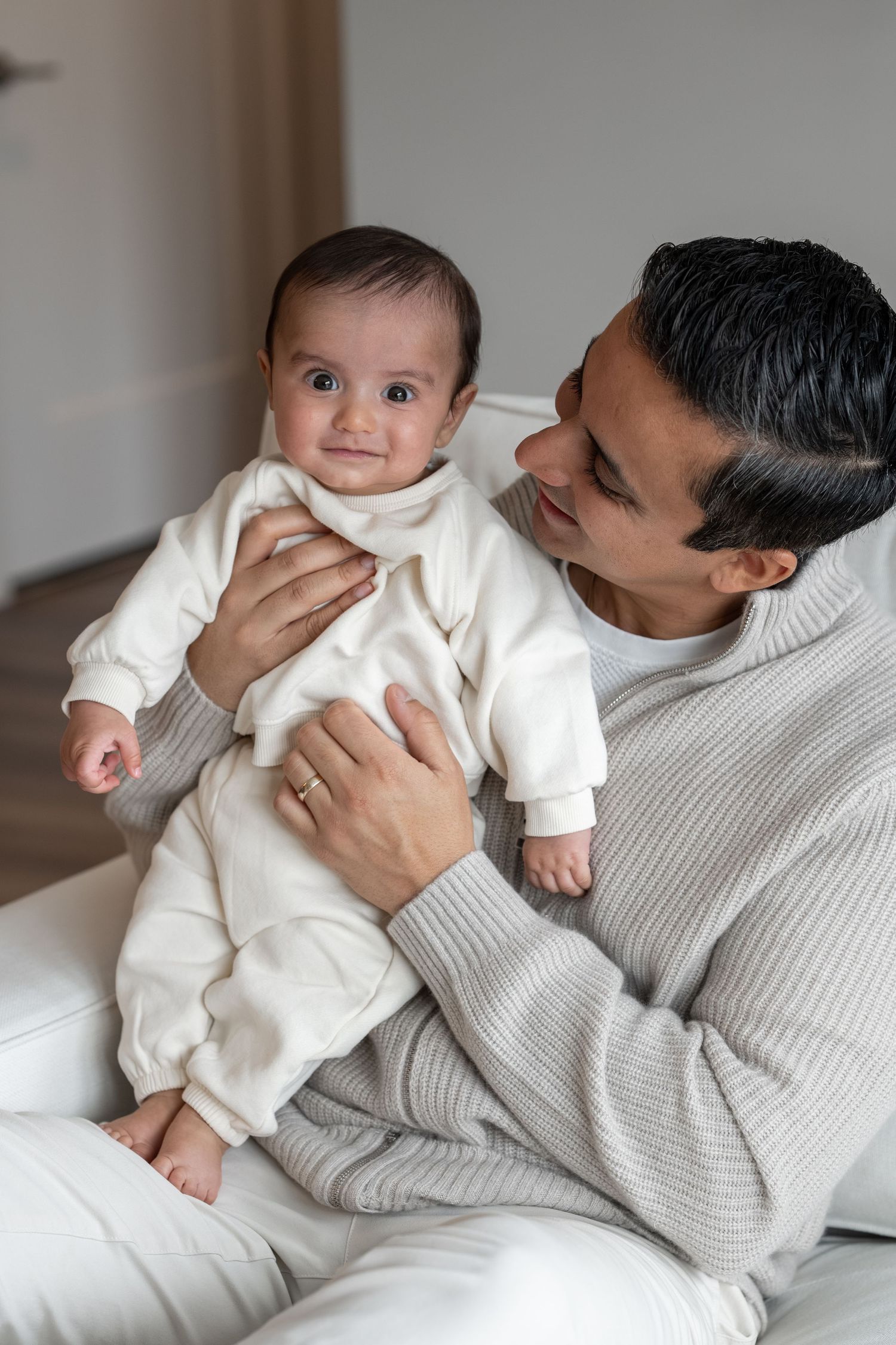 Loving father in gray sweater holds smiling baby dressed in white outfit on white sofa.