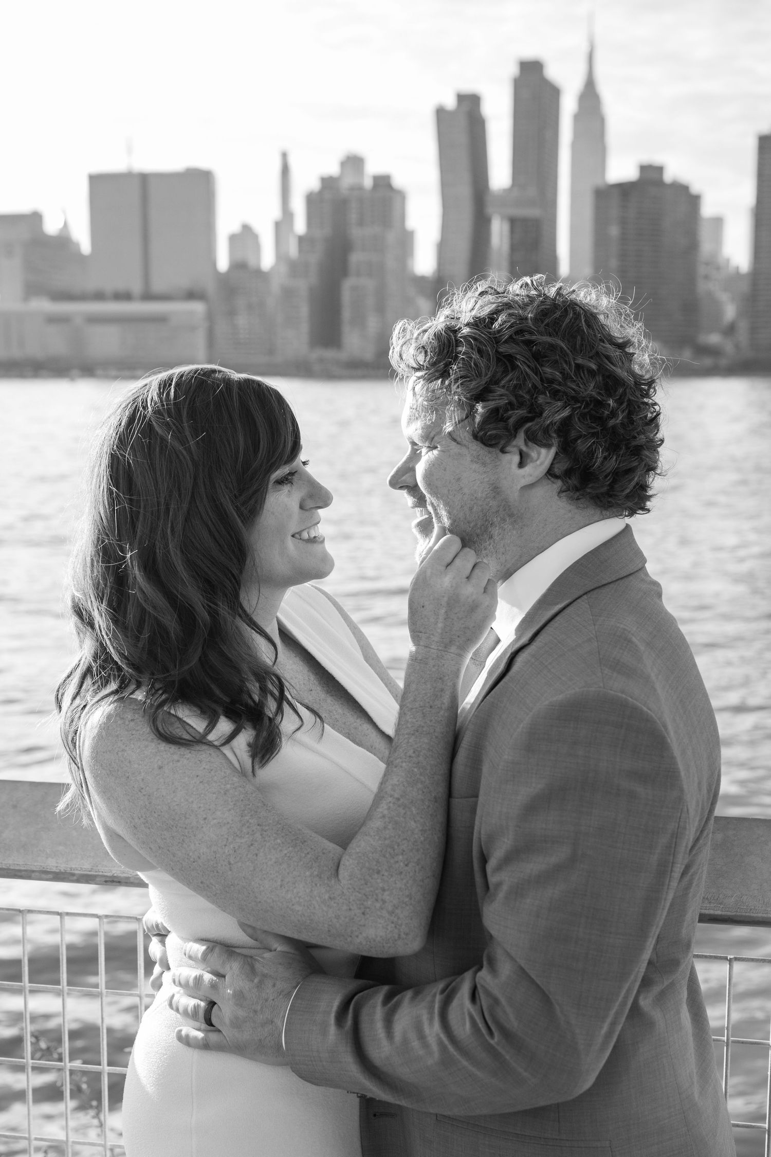 Romantic wedding couple embracing with New York City skyline visible across the water in black and white.