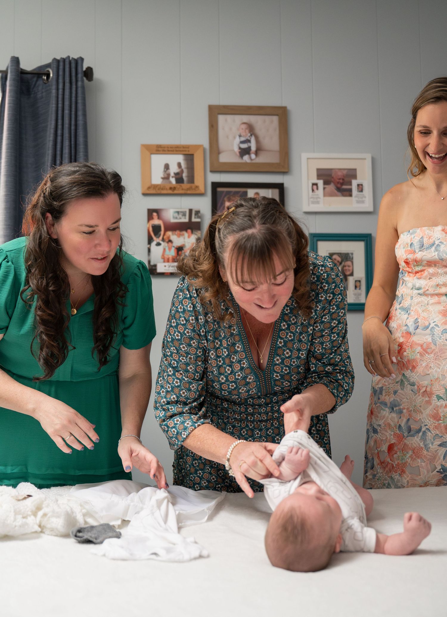 Three women gather around a changing table to care for an infant in a room with framed photos on the wall.