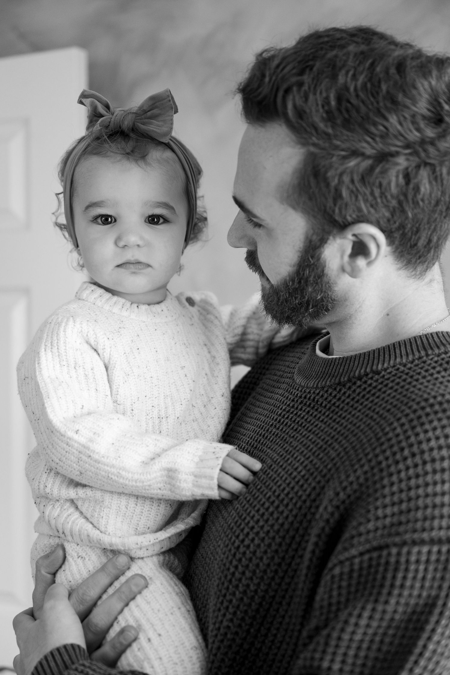 Black and white portrait of parent in sweater holding baby wearing bow and knit outfit.