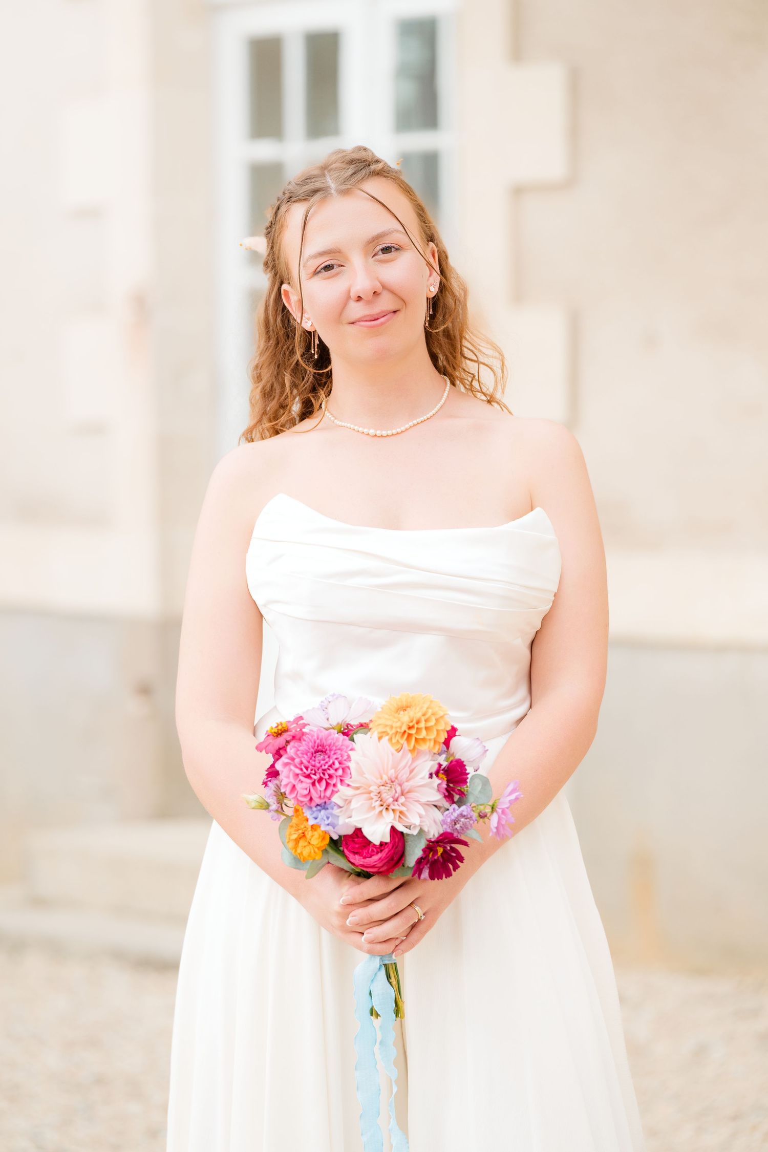A bride in a white strapless dress holds a vibrant bouquet of pink and yellow flowers against a light-colored building.