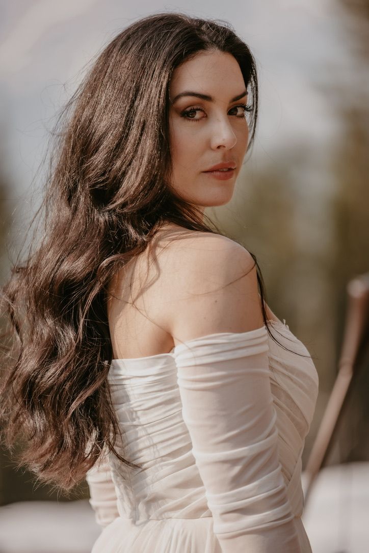 Elegant woman in white off-shoulder dress poses with long dark hair against blurred background.
