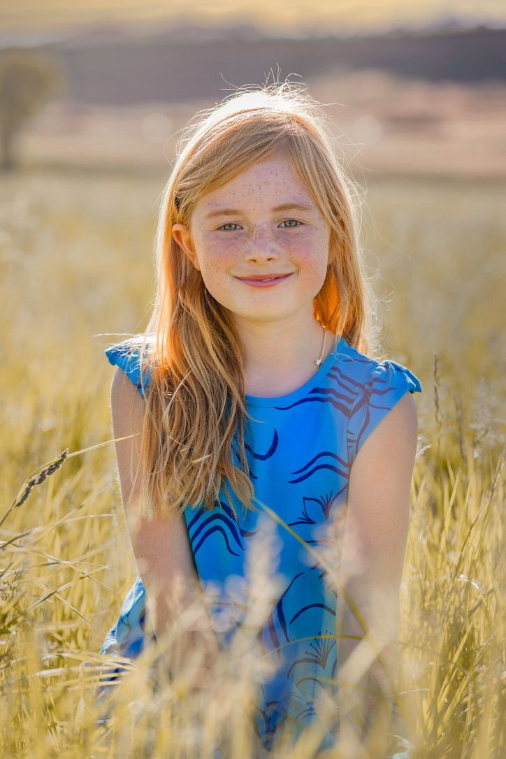 A young person in a bright blue dress smiles while standing in a sunlit field of tall grass.