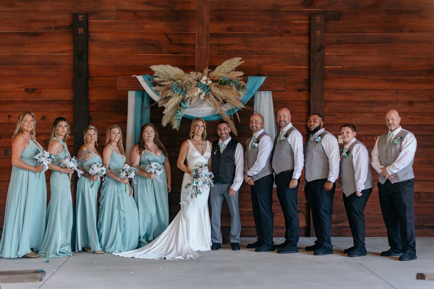 Wedding party lines up against rustic wooden wall wearing turquoise dresses and white shirts.