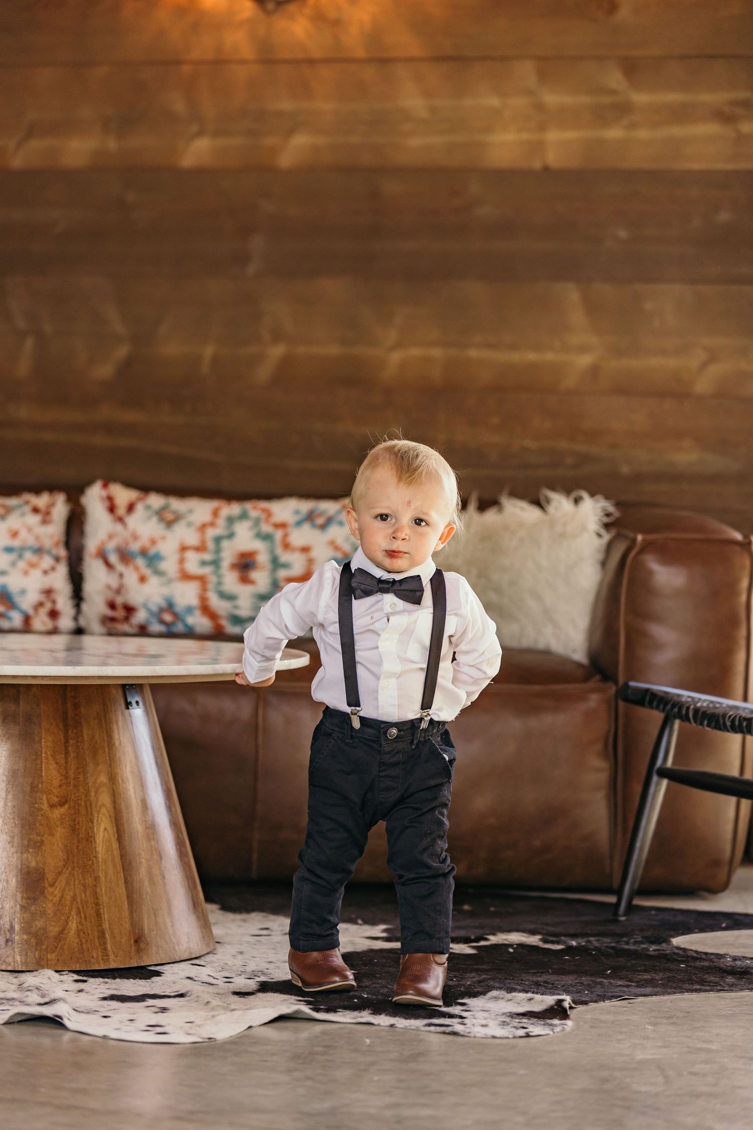 Small child in formal wear with suspenders standing near leather furniture in rustic indoor setting.
