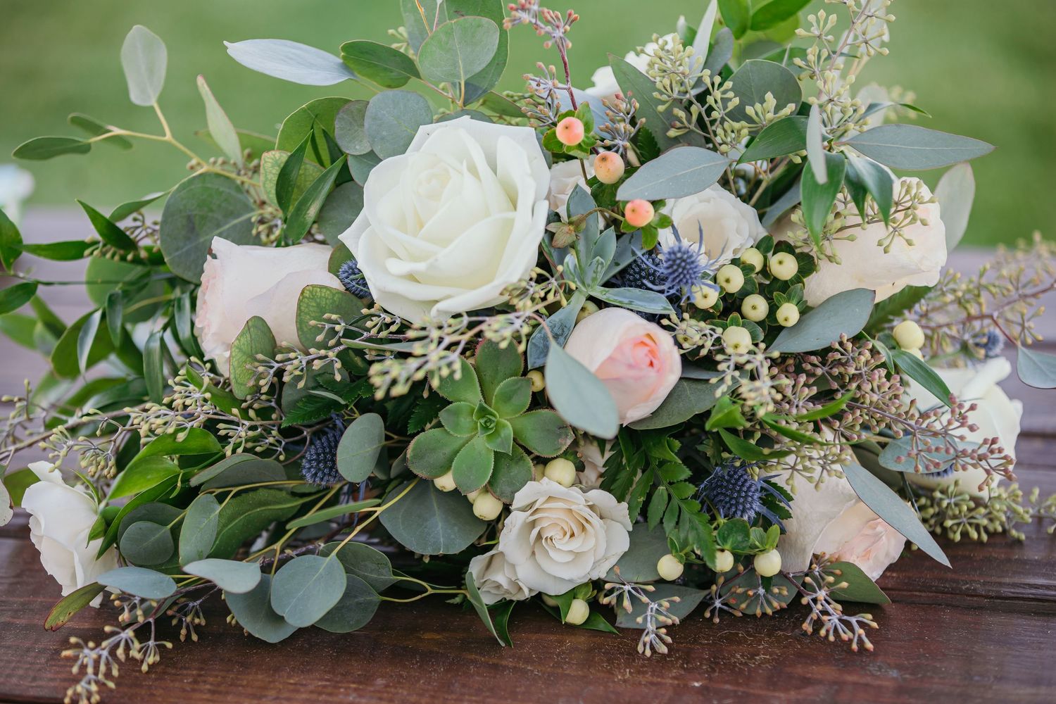 Wedding floral arrangement featuring white roses, eucalyptus leaves, and small berries on a wooden surface.