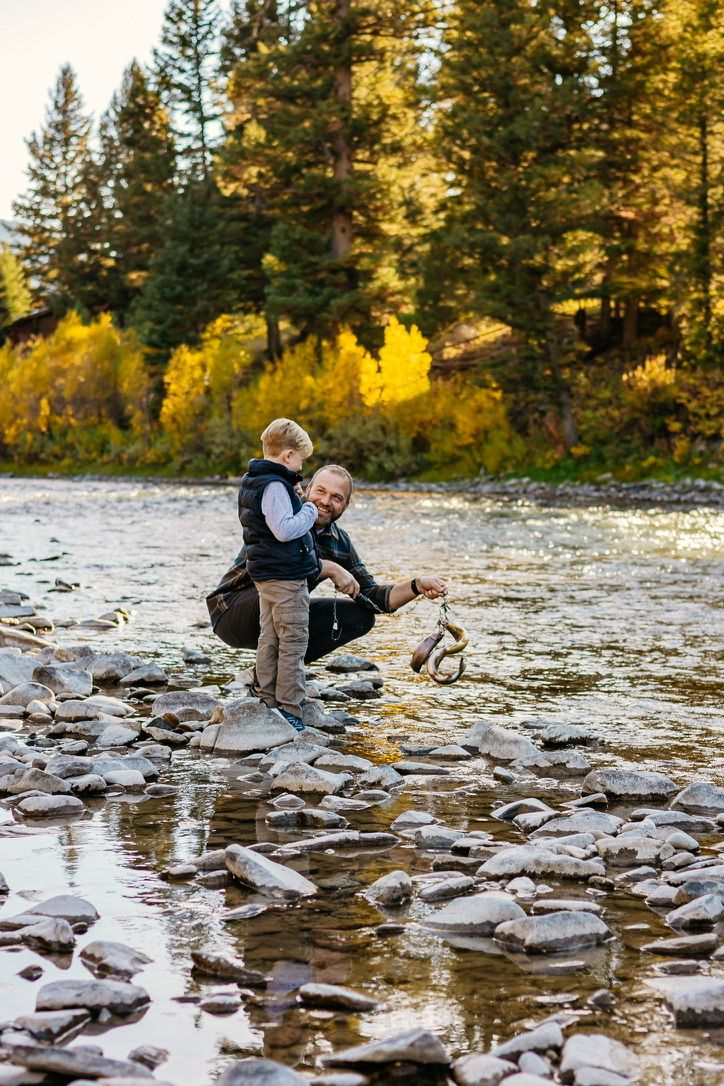 A parent and child explore rocky riverbanks together during golden hour in a forest setting.