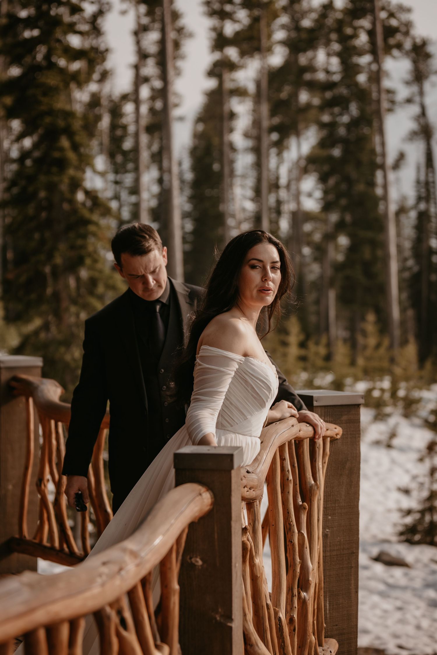 Winter wedding couple poses on rustic wooden balcony with snow-covered forest backdrop.