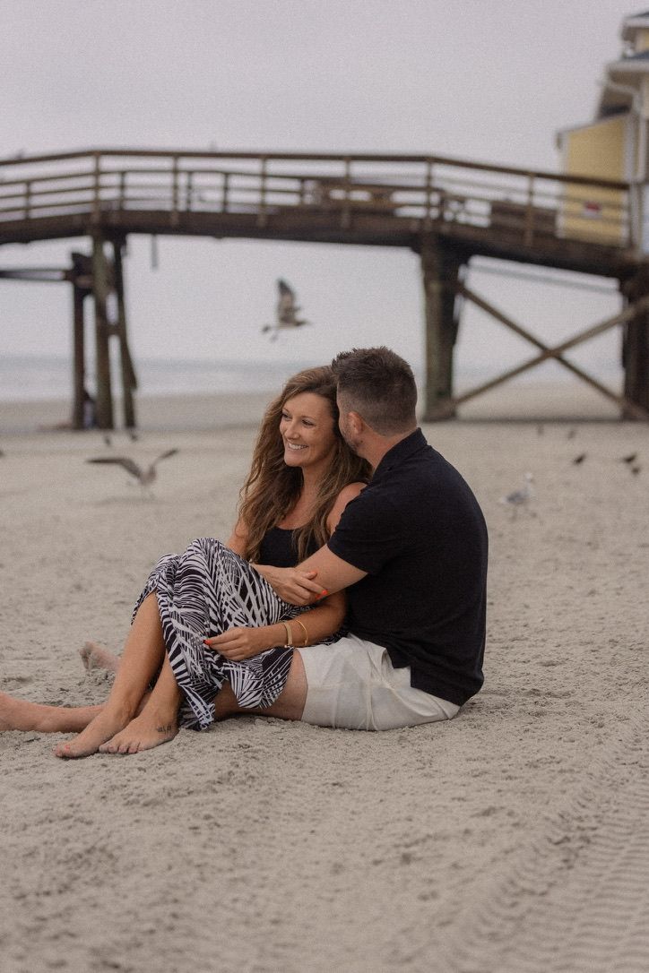 Couple lying on sandy beach near wooden pier during romantic photoshoot.