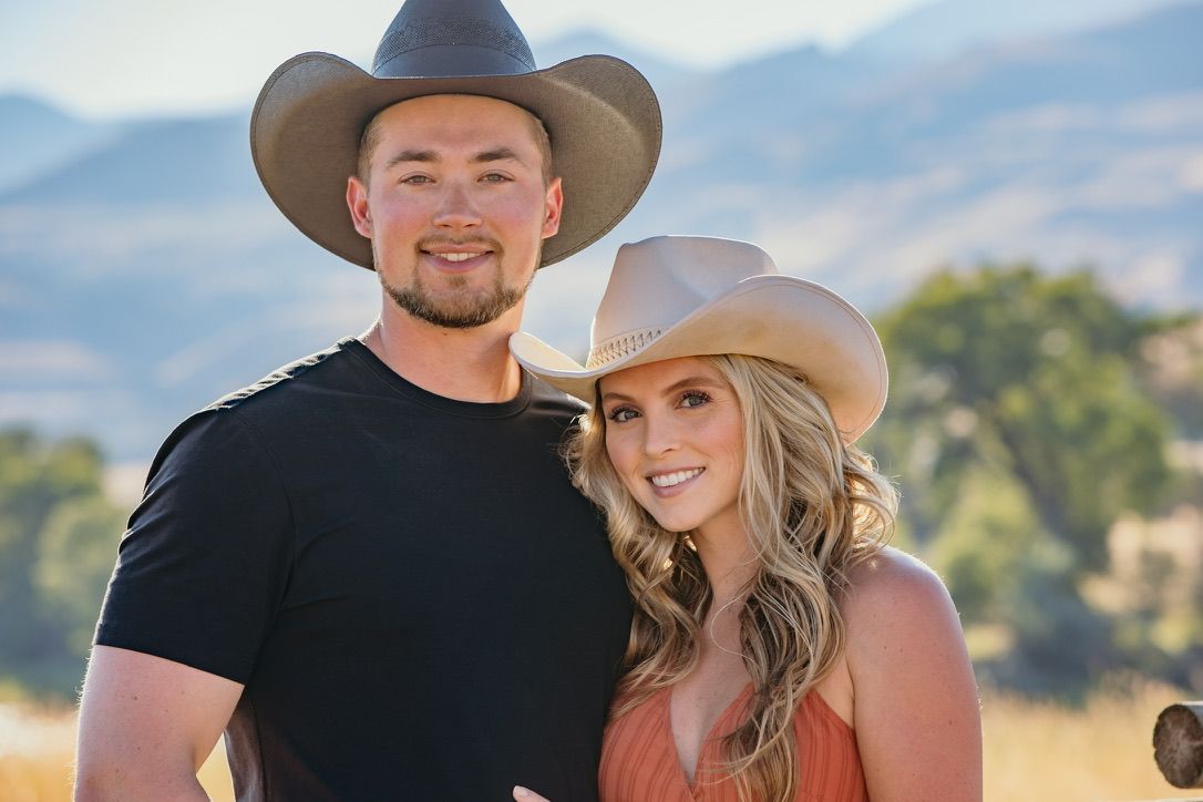 Couple walking through field with mountains and cowboy hat in background.