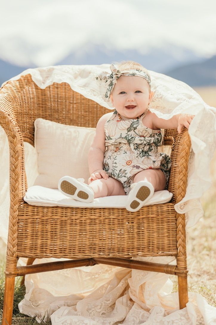 Baby wearing floral outfit sits in wicker chair with white lace backdrop and cushion.