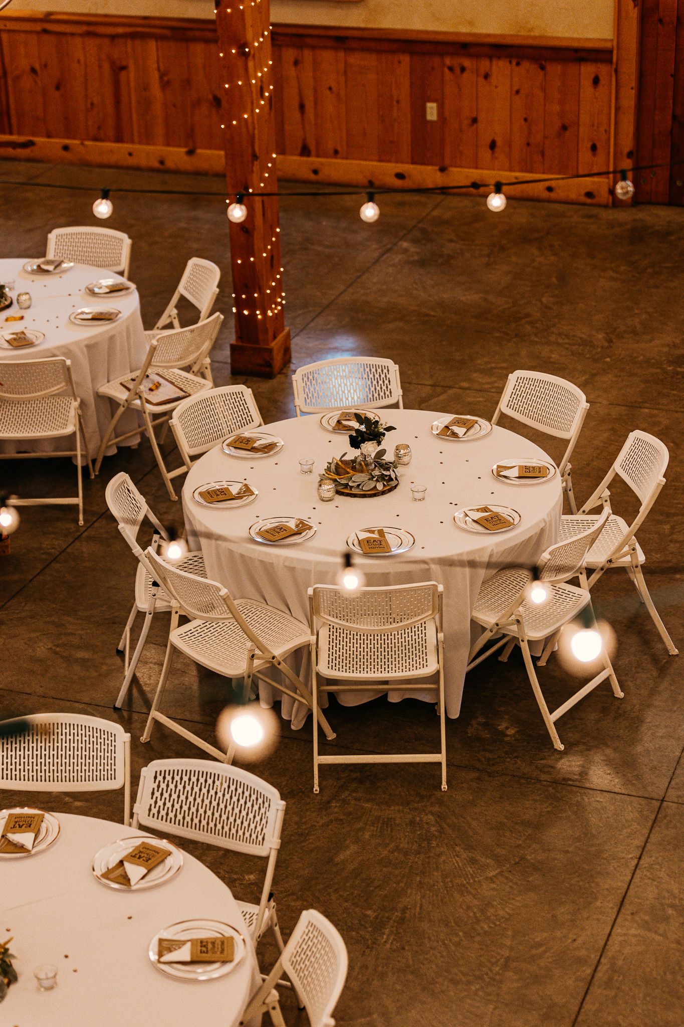 Round reception tables with white tablecloths and chairs set up under string lights in a rustic barn venue.