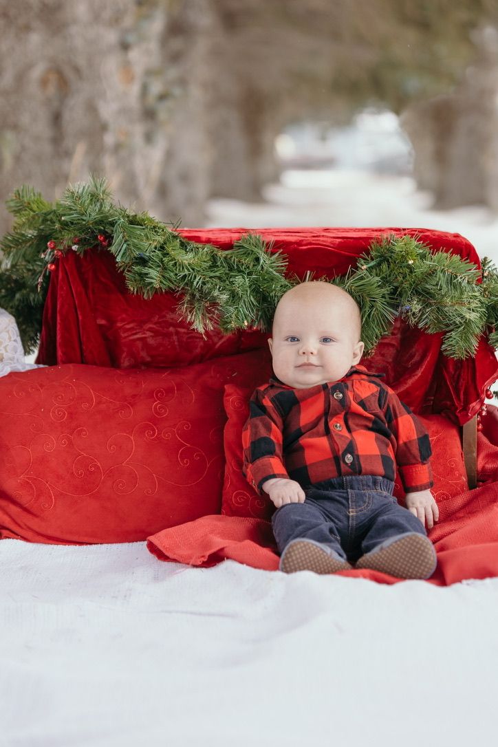 Baby in red plaid shirt sits among holiday decorations with evergreen garland and red velvet.