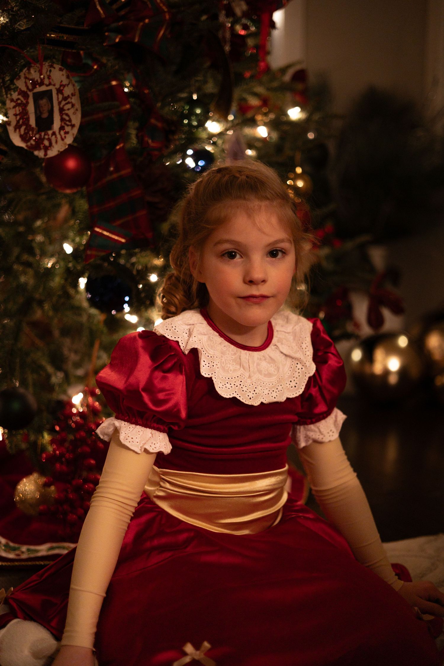 Young girl in red velvet Christmas dress sitting in front of decorated holiday tree with glowing lights.