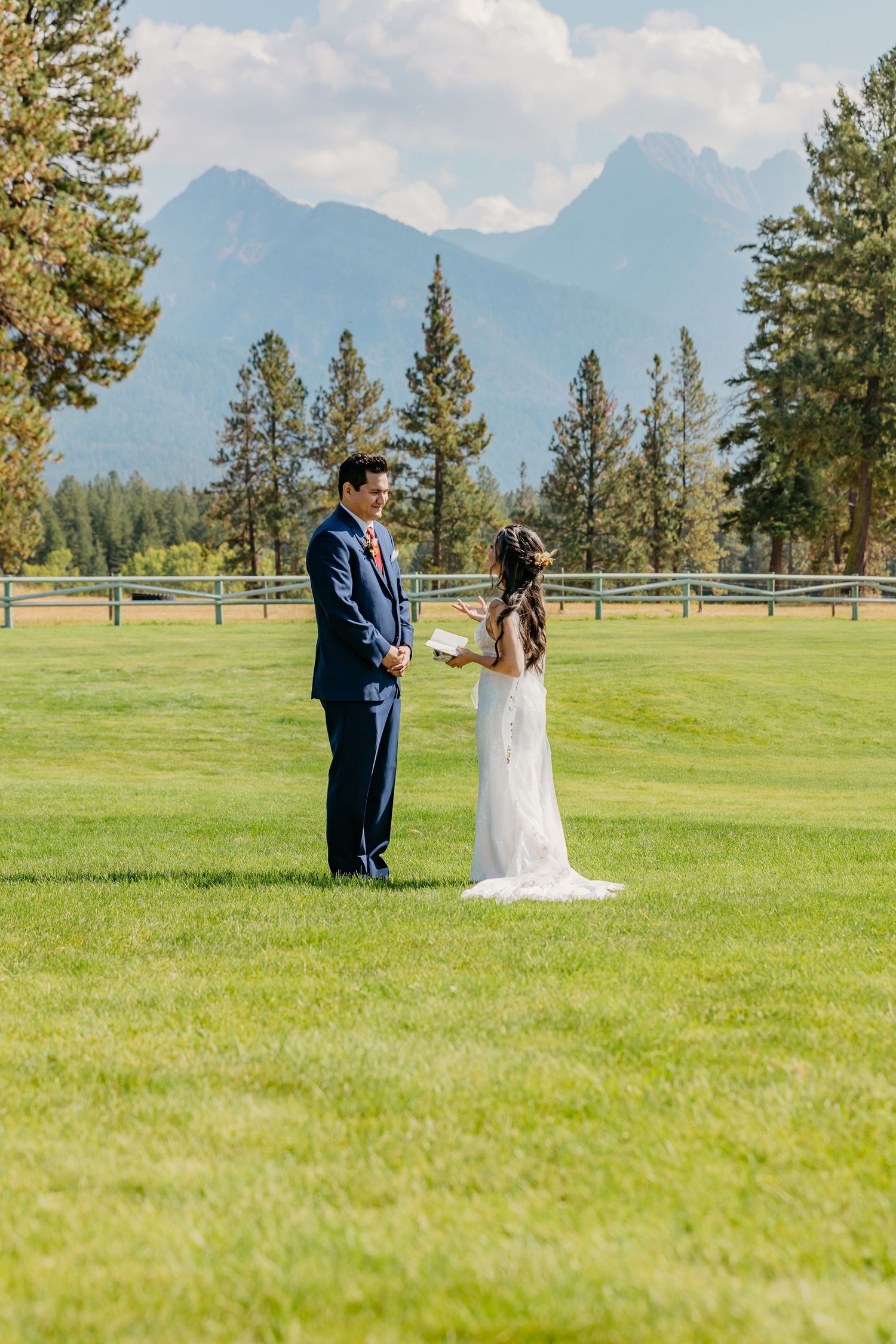 A couple stands together on a lush green lawn with majestic mountain peaks and evergreen trees in the background.