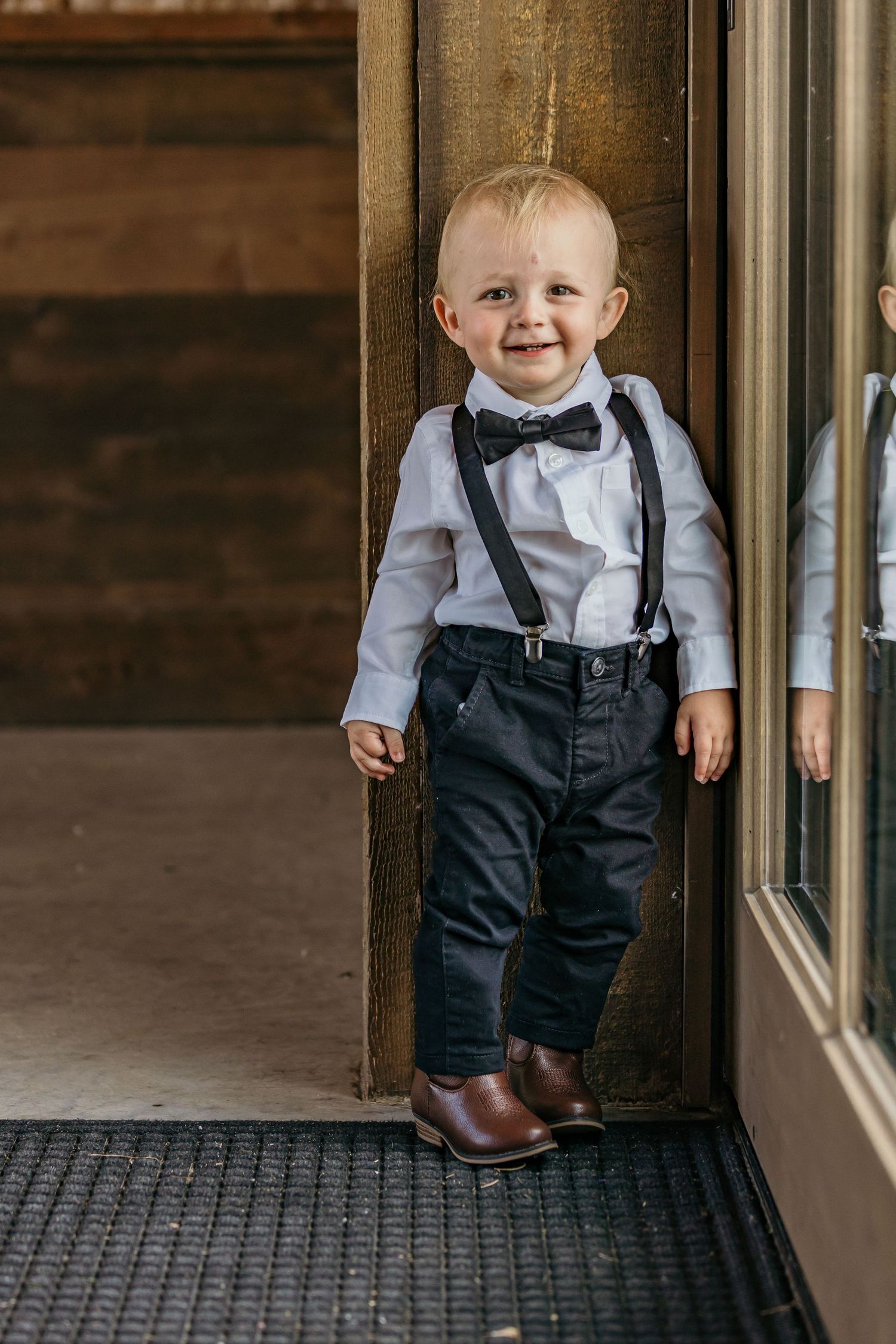 Small child wearing formal attire with suspenders and bow tie standing by a window.