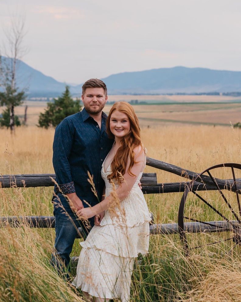 Couple poses playfully by rustic wooden fence in golden field with mountains in background during sunset.