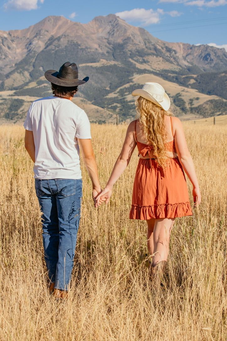 Couple walking hand in hand through tall grass with mountain backdrop.