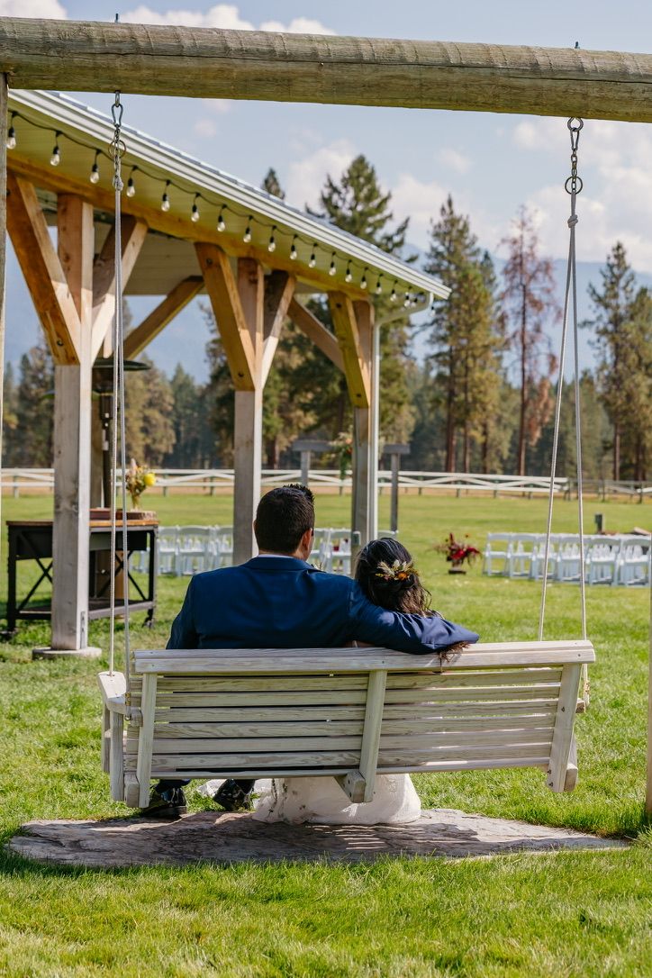 Couple sitting on a wooden swing bench at an outdoor wedding venue with string lights overhead.