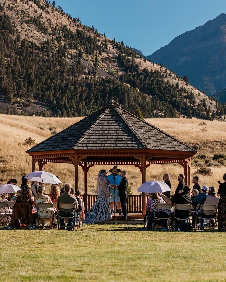 A mountain wedding ceremony takes place under a wooden gazebo with guests seated on white chairs.