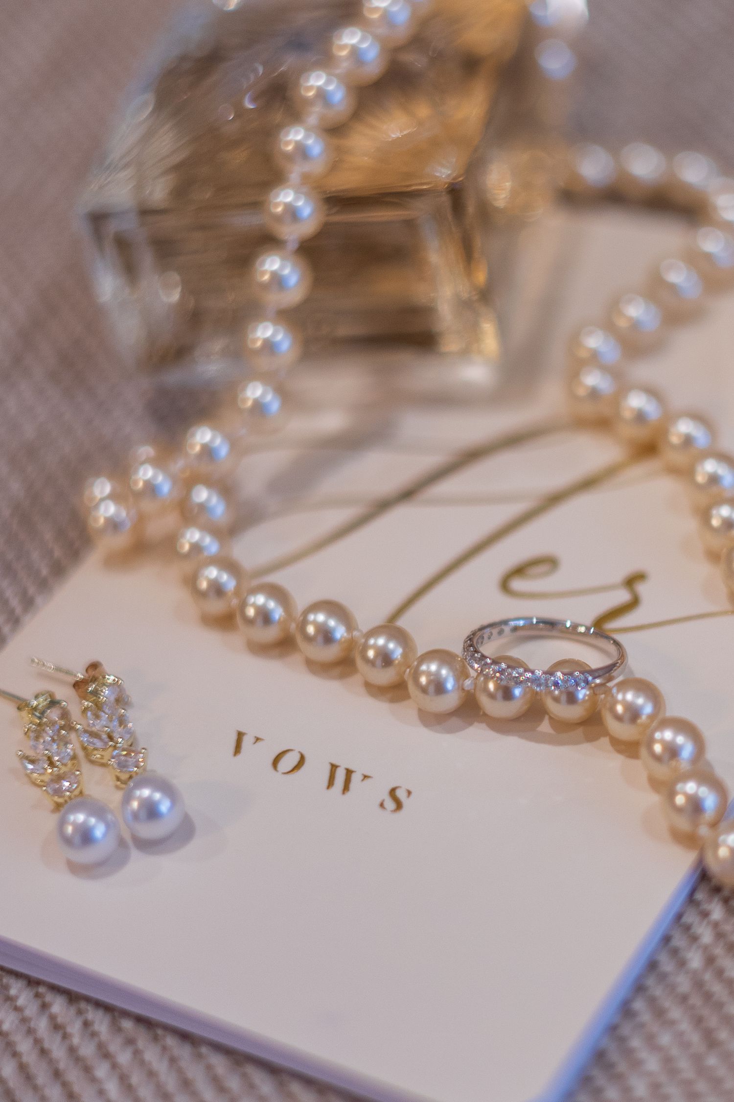 Pearl necklace and diamond ring resting on a vows book with pearl earrings and perfume bottle for a wedding flat lay.