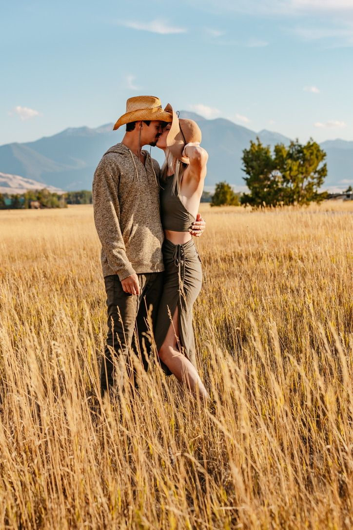 Couple embracing in golden wheat field with mountains and blue sky in background.