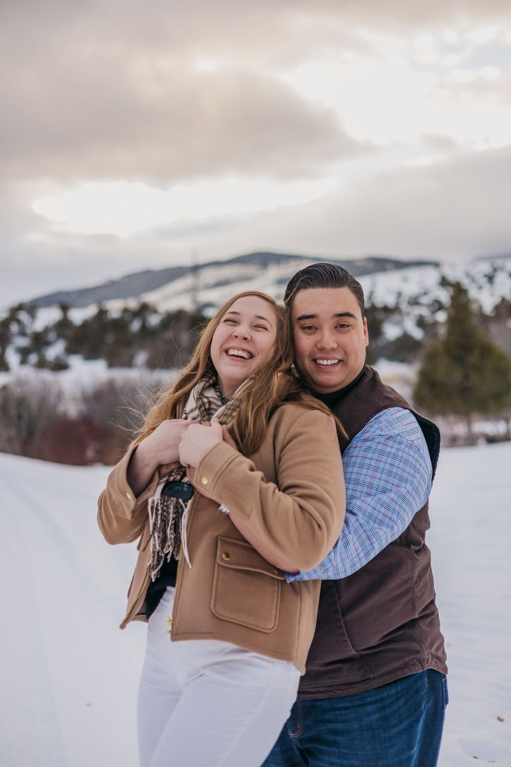 A couple embraces and laughs together in a snowy winter landscape with mountains in the background.