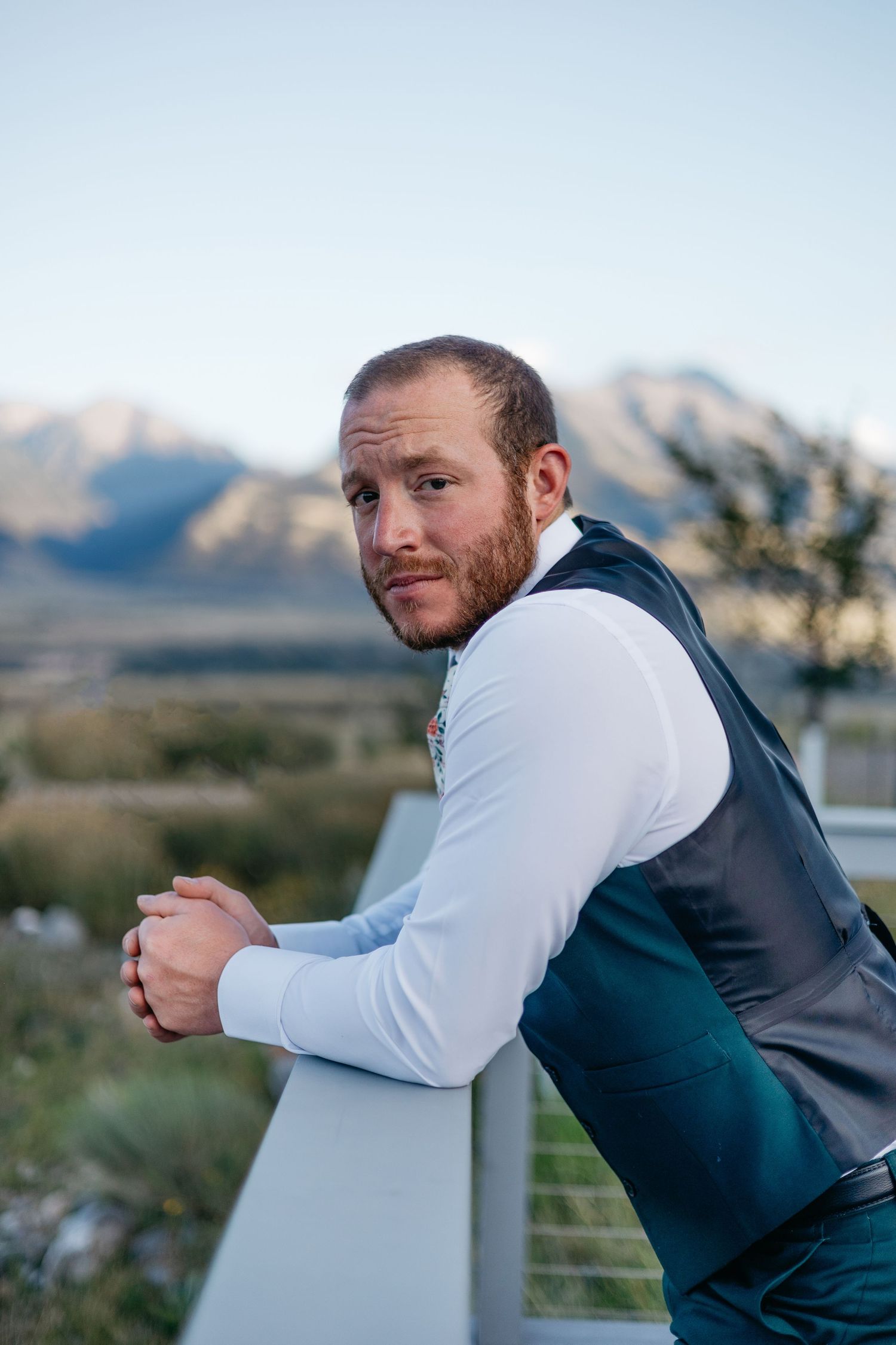 Person in formal attire leaning on outdoor railing with mountains in background during golden hour.