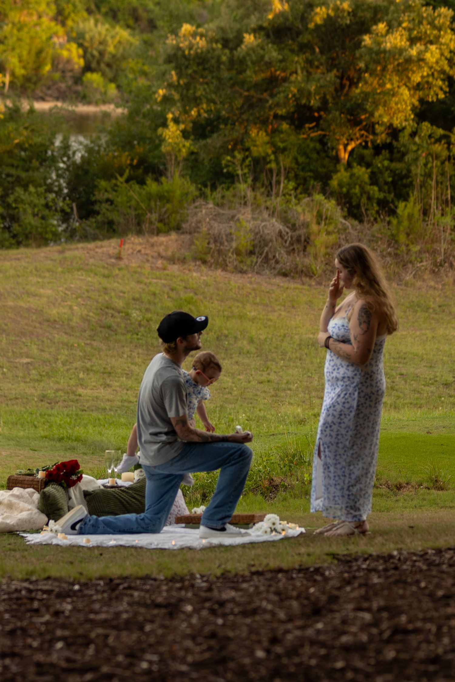 Man kneeling to propose to woman in floral dress during outdoor picnic with child present on grass.