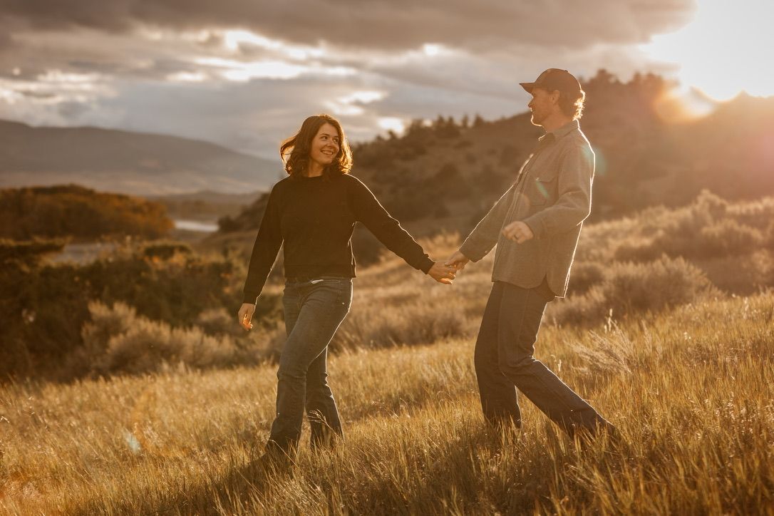 Couple running through golden field at sunset with mountains in background.