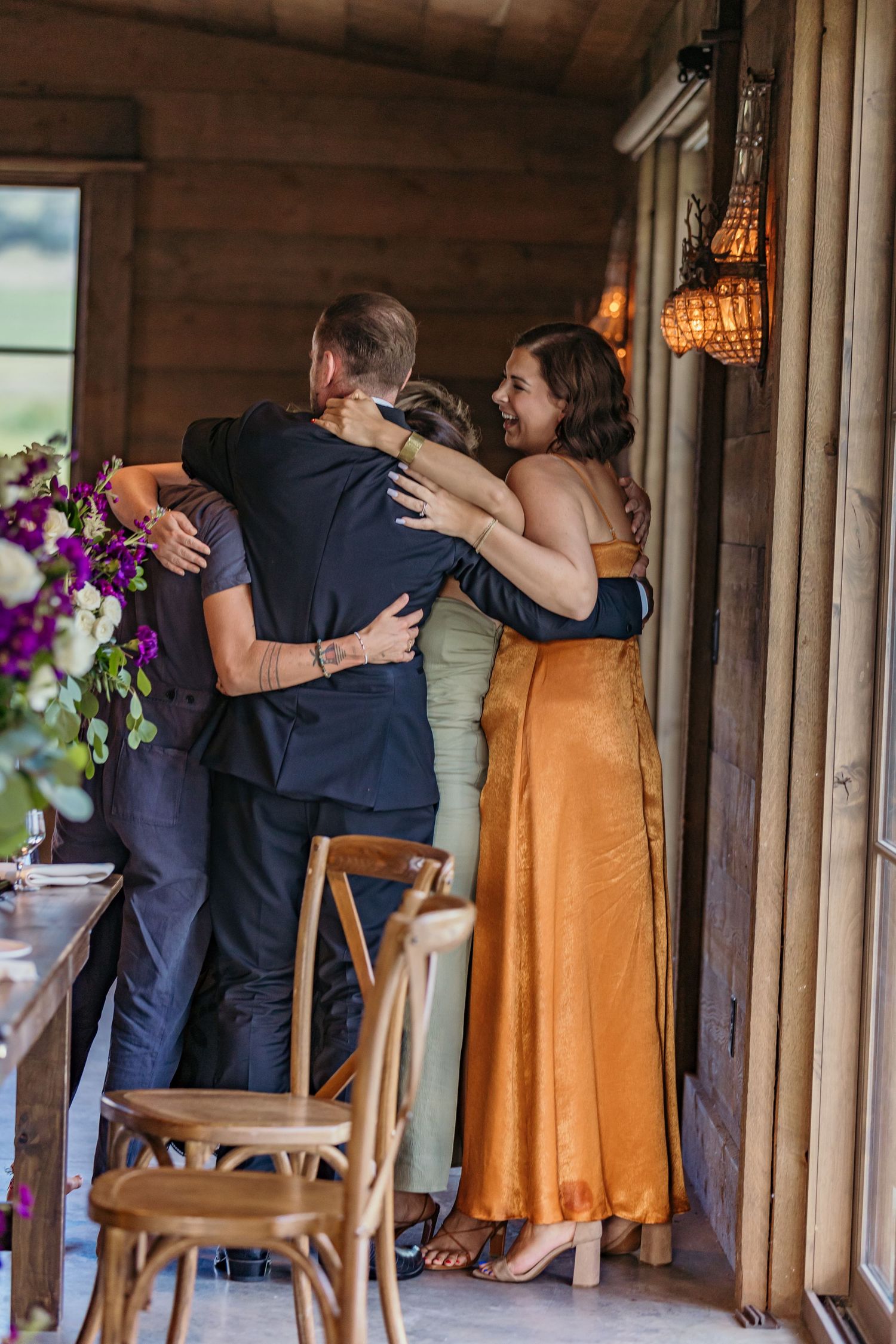 Wedding party embraces in rustic venue with wooden columns and warm lighting.
