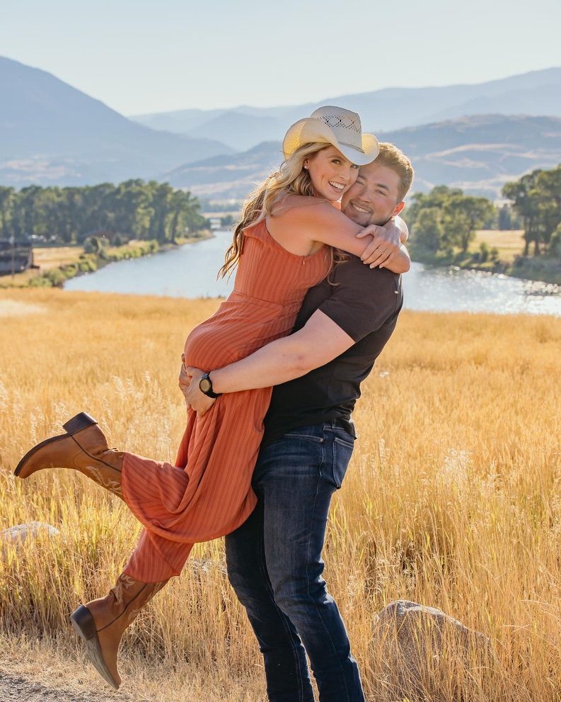 Joyful couple embracing in golden field with mountain lake vista behind them.