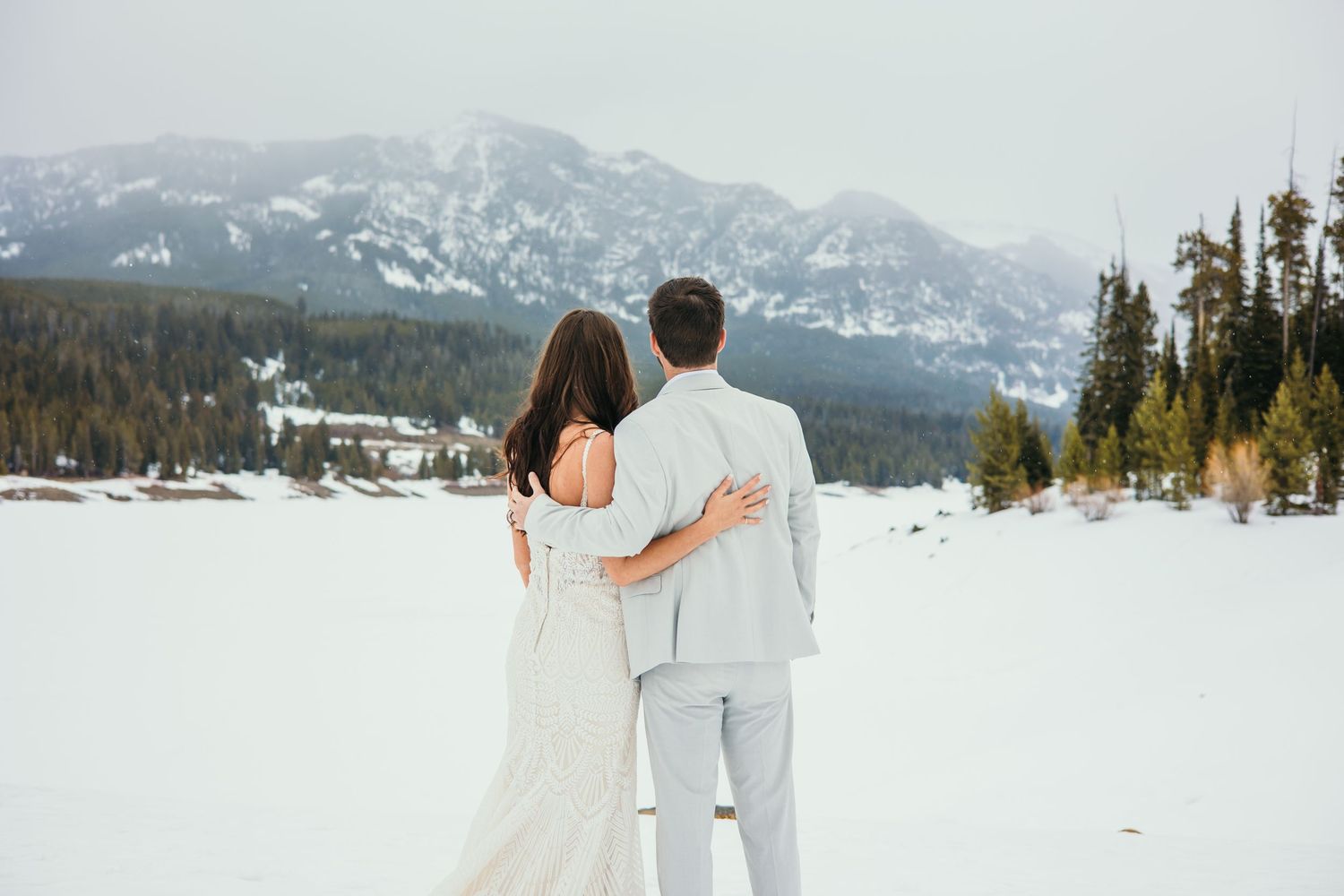 A couple in wedding attire embrace while looking at snow-covered mountains in a scenic winter landscape.