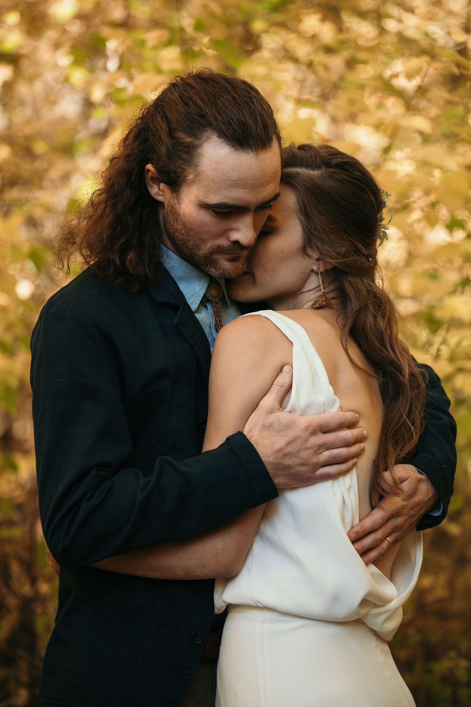 Romantic autumn wedding moment with couple embracing outdoors against golden fall foliage background.