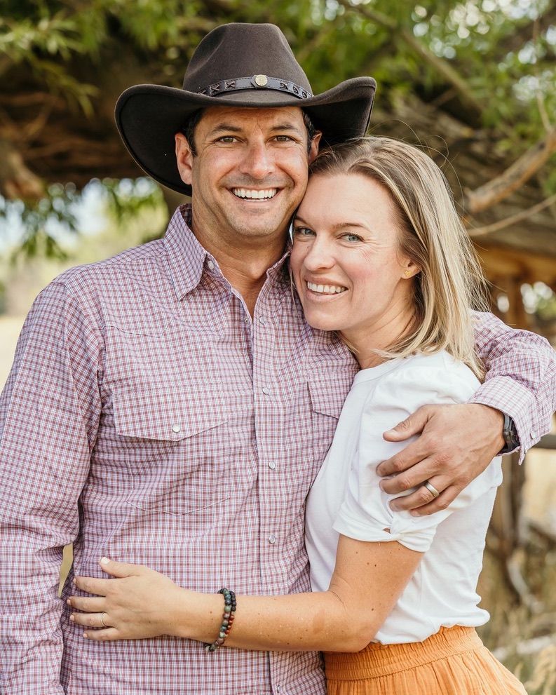 A couple in western attire share an embrace while posing outdoors in casual country style clothing.