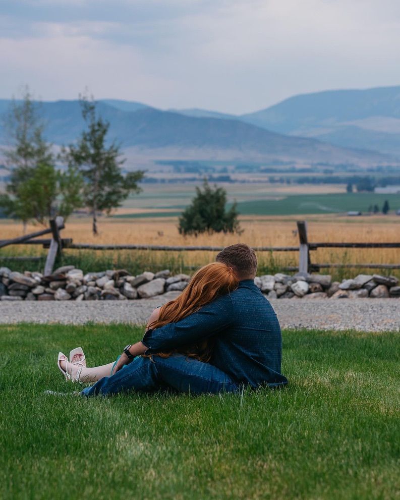 Person in denim sits peacefully on grass overlooking mountain valley landscape.