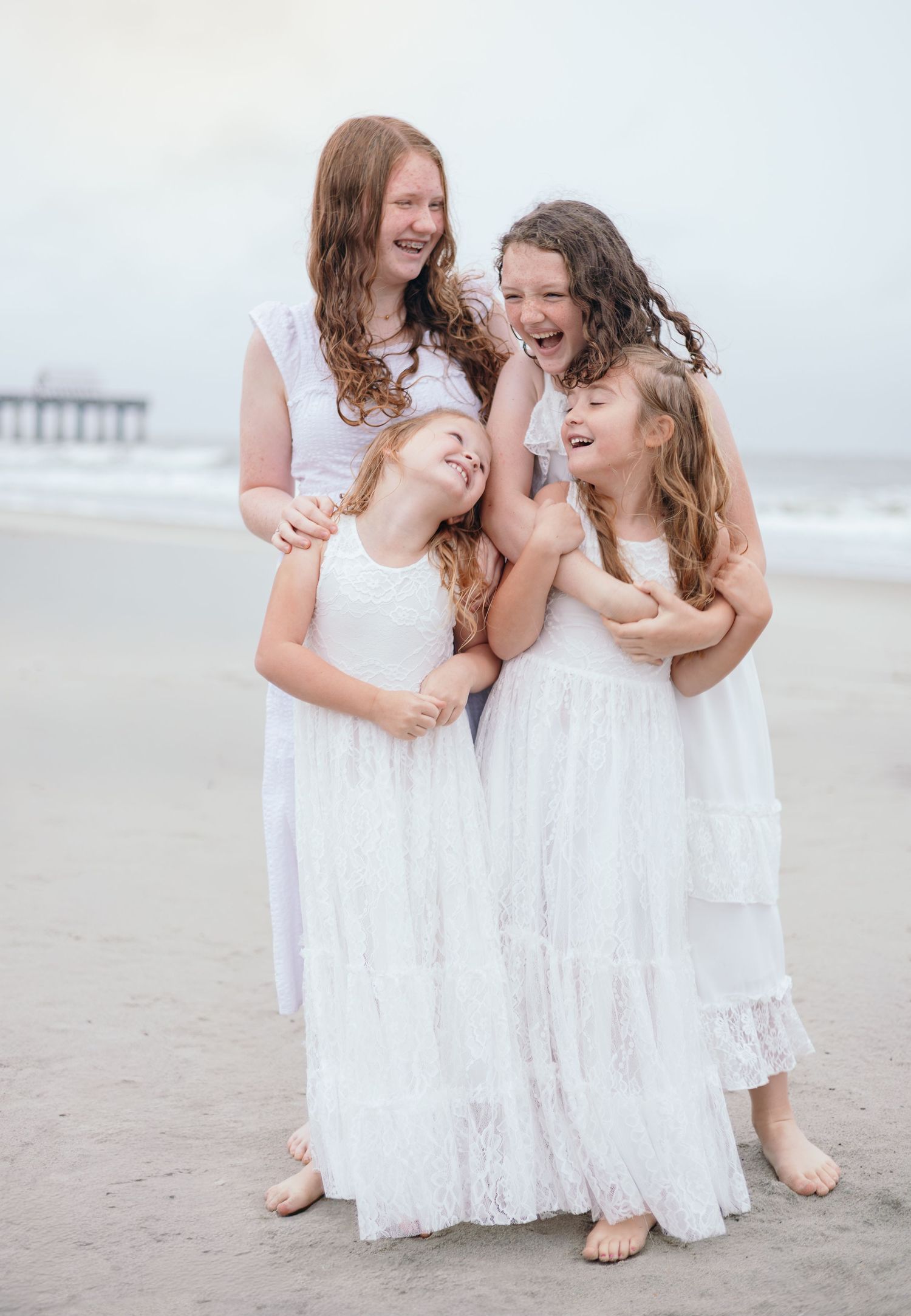 A group in flowing white dresses shares joyful moments together on a misty beach near a pier.
