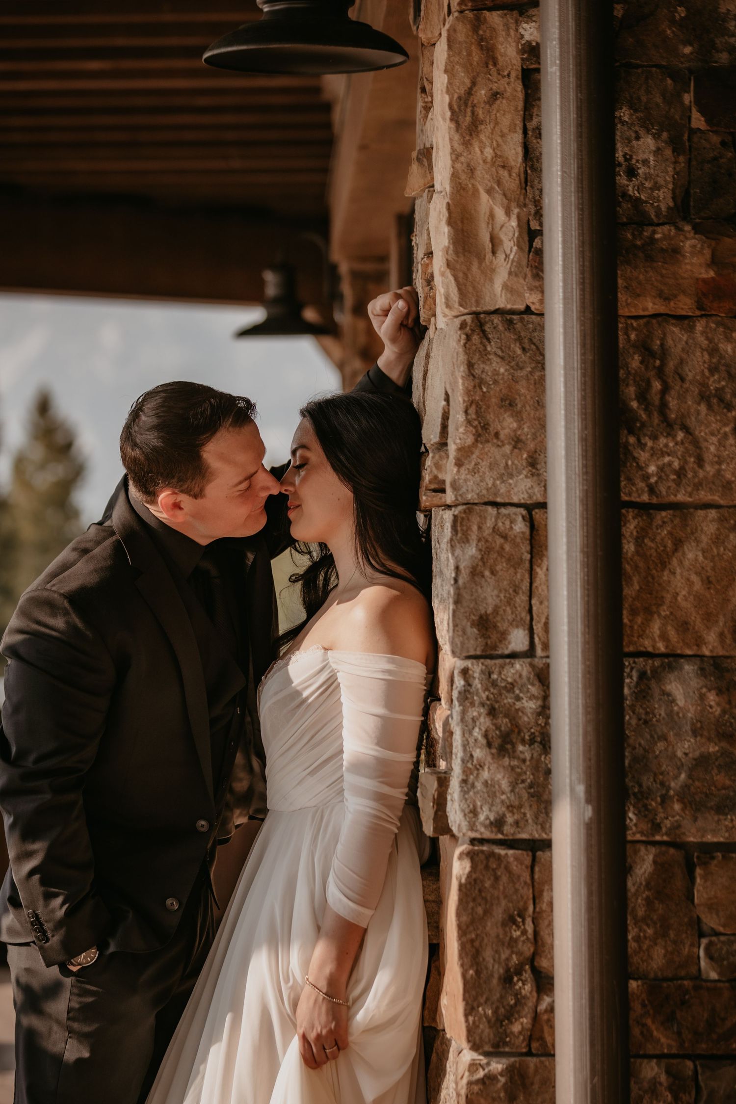 A romantic wedding photo at sunset showing a couple in a tender moment against a rustic stone wall.