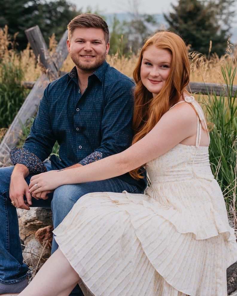 A couple poses casually on a log in a natural setting wearing blue and white outfits.