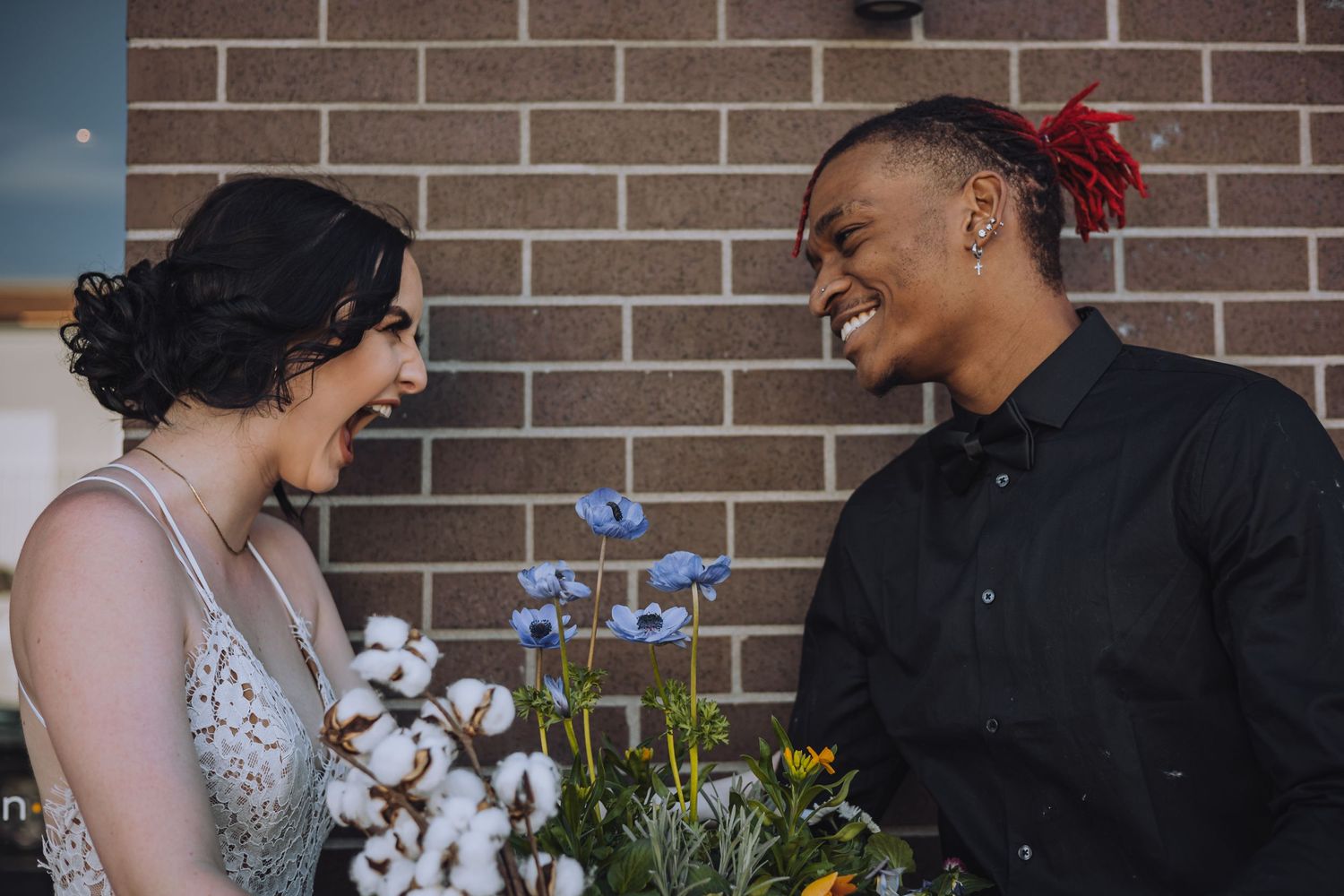 Two people share a happy moment together in front of a brick wall with cotton plants and purple flowers.