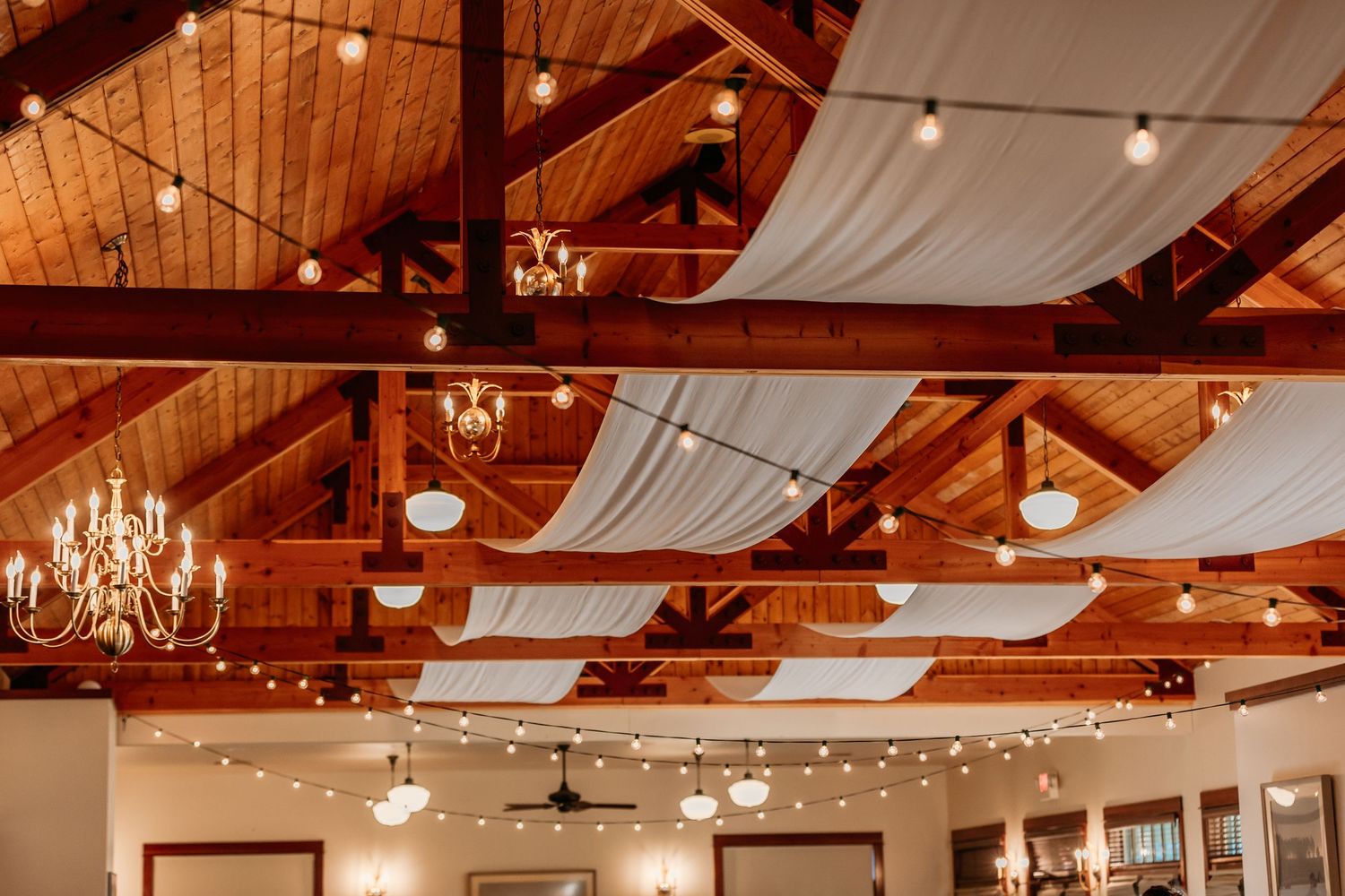Wedding reception hall with exposed wooden beams, chandeliers and string lights.