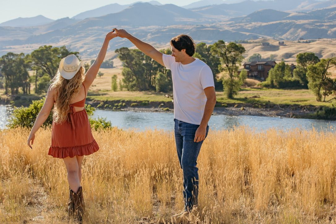 A couple dances playfully in a field with mountains and scenic landscape behind them.