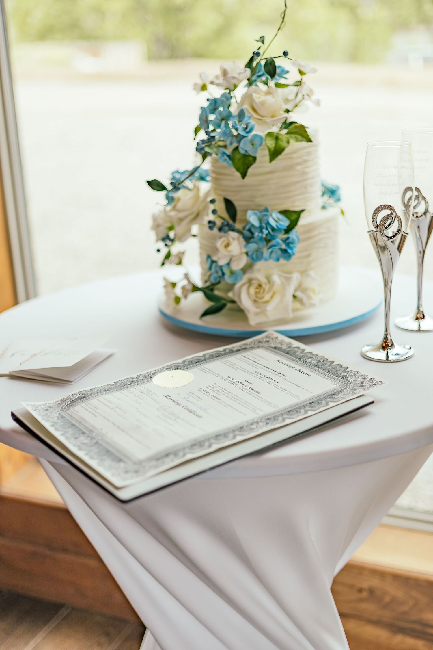 White wedding cake decorated with blue flowers next to silver document holder.
