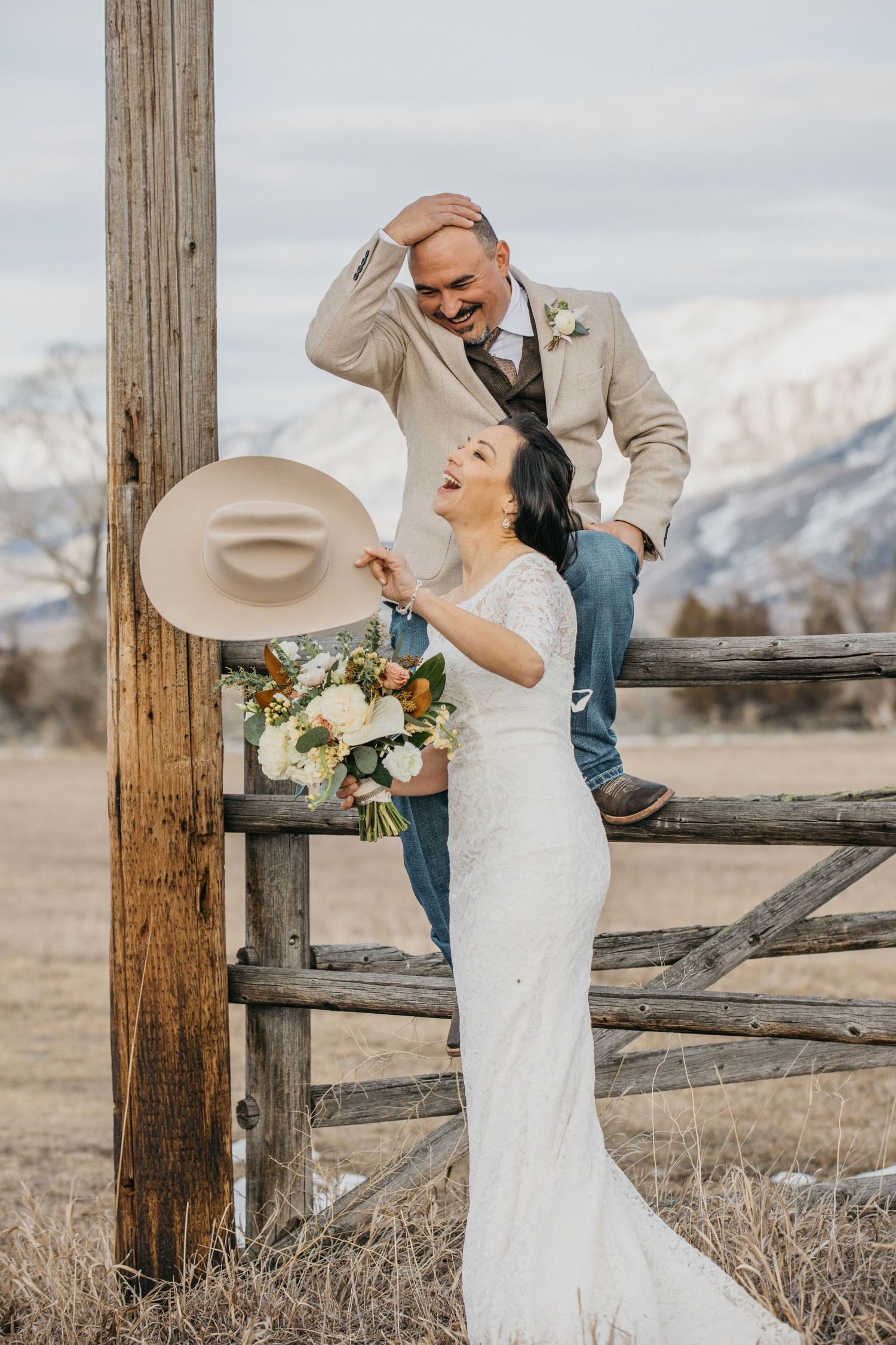 Romantic wedding couple poses by wooden fence with snow-capped mountains in background.