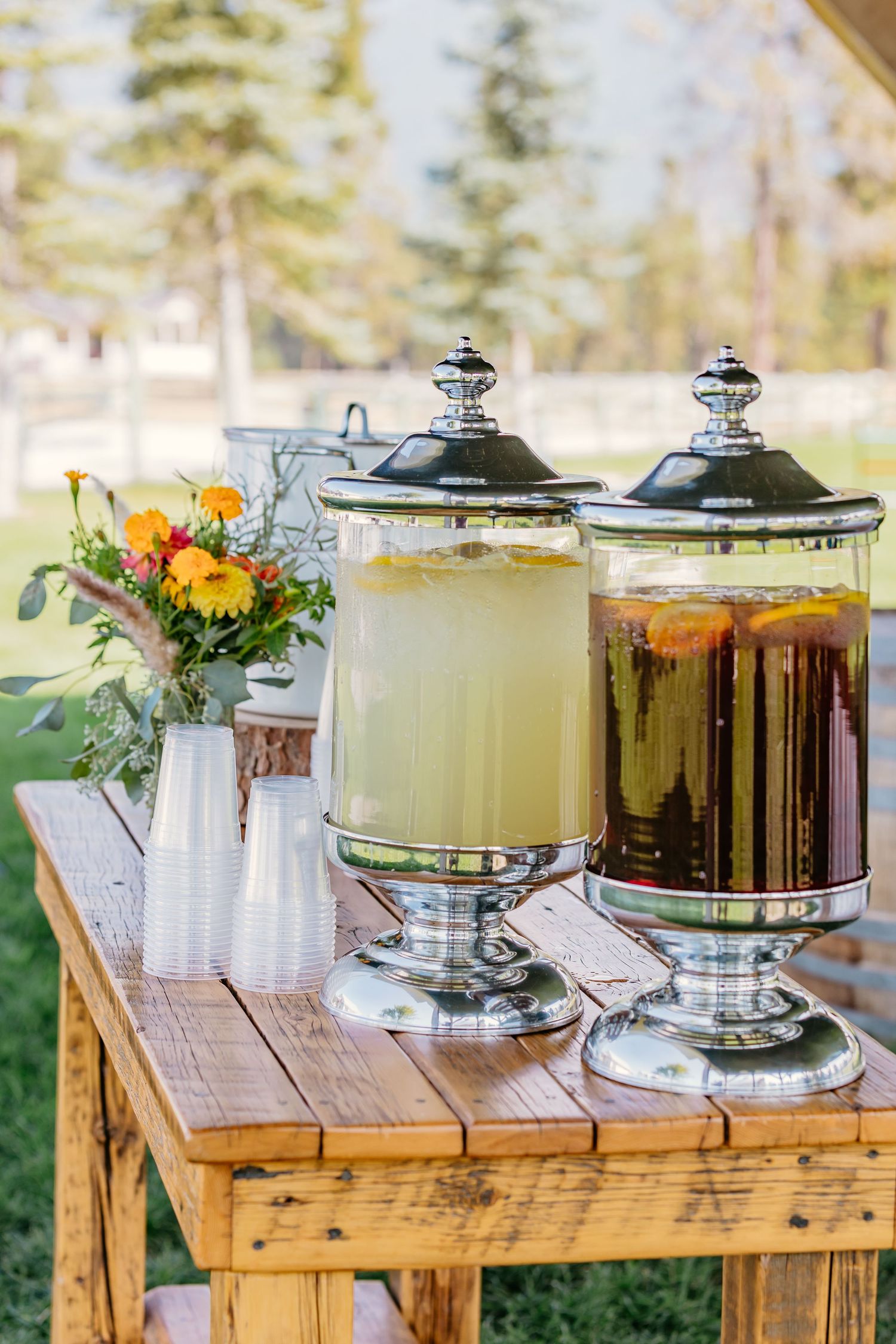 Rustic wooden table with beverage dispensers containing refreshing drinks and floral decorations for an outdoor event.