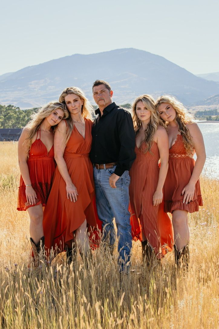 A group in matching rust-colored dresses poses in a golden field with mountains behind.