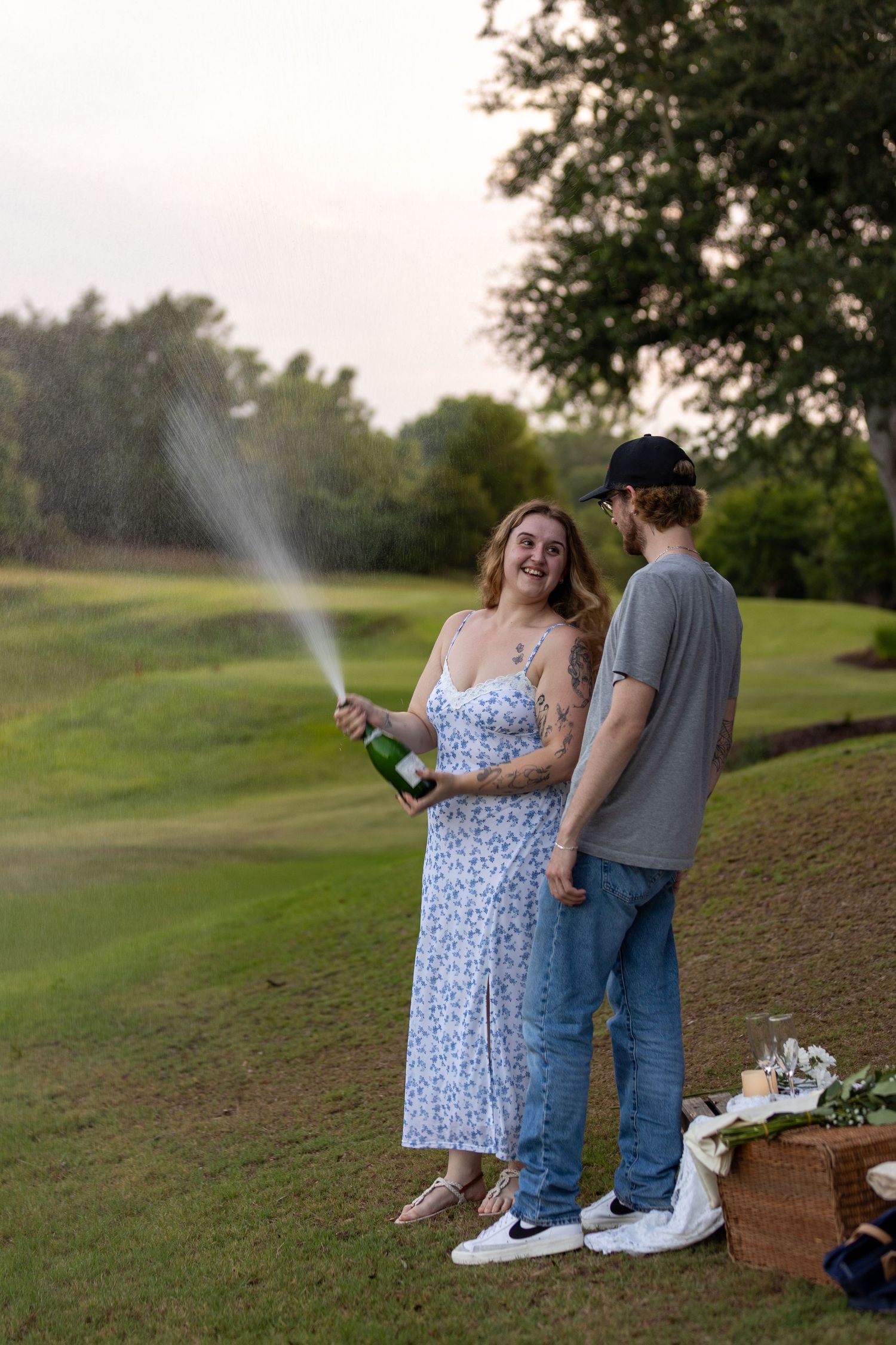 Couple celebrating on golf course, woman in floral dress spraying champagne bottle, man in gray shirt and cap beside her.