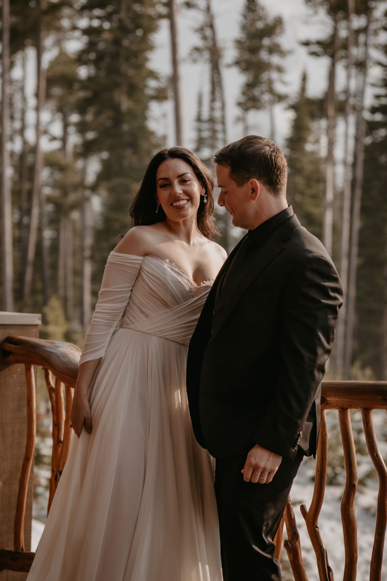 Romantic wedding couple sharing a moment on a wooden deck with pine trees in the background.