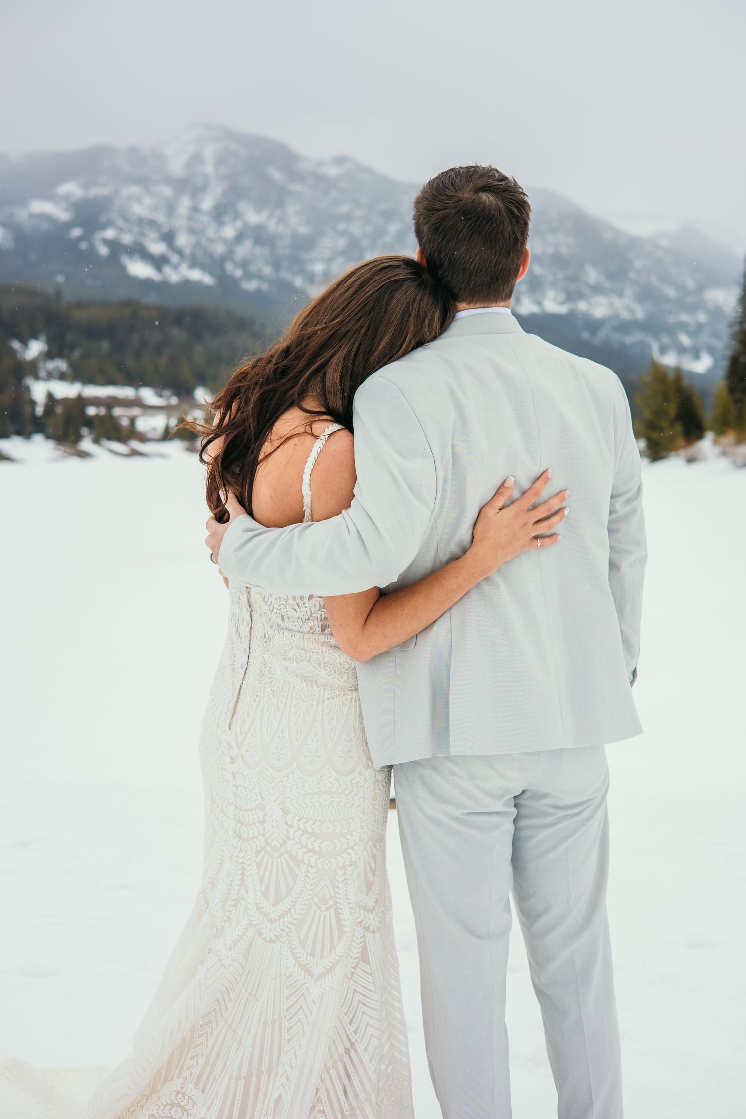 A couple in white wedding attire stands together against a snowy mountain landscape.