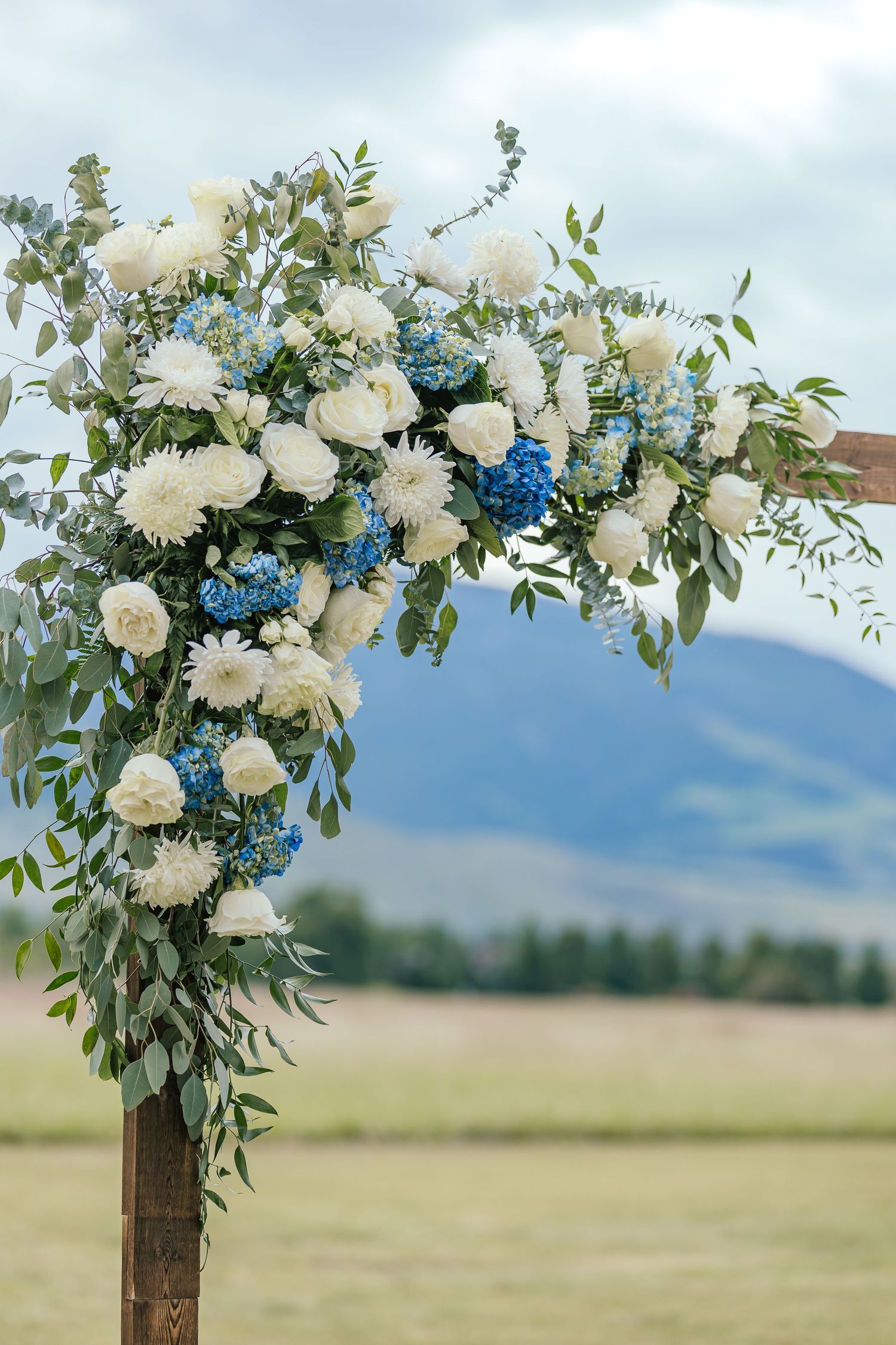 White and blue floral wedding arch arrangement against mountain landscape.